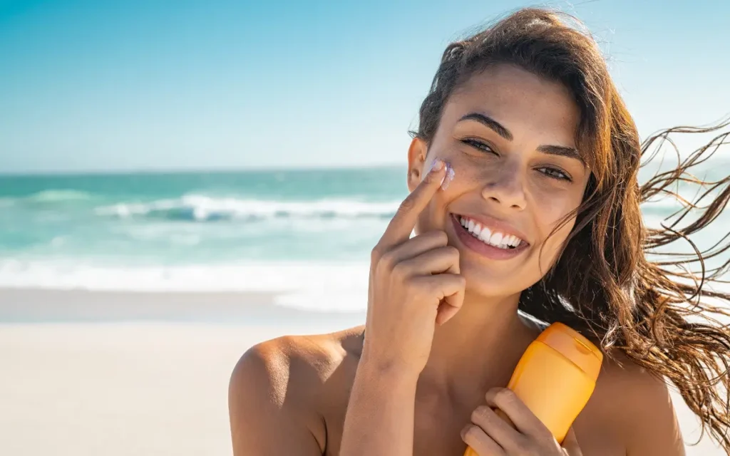 Protetor Solar: Cuidados Primordiais Para a Pele do Rosto - Mulher na praia segurando um pote de protetor solar e passando no rosto