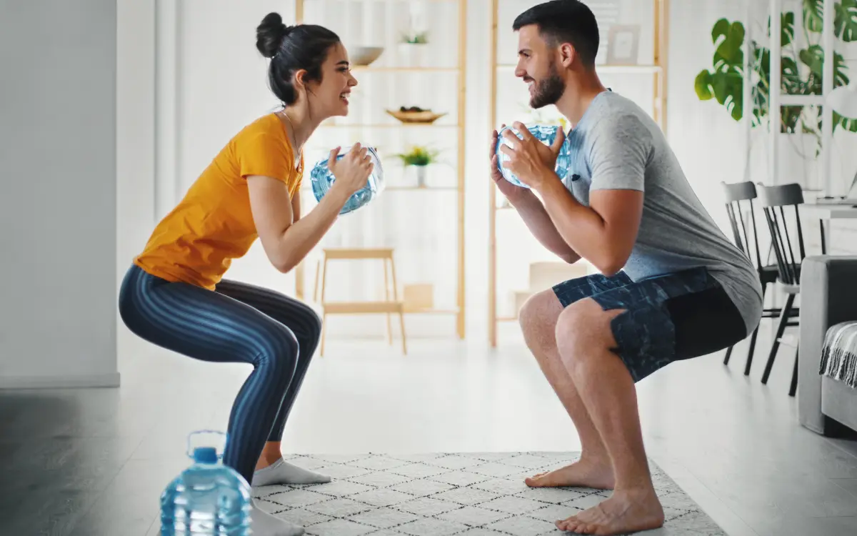 Treino em Casa: 3 Dicas Básicas Para Emagrecer Com Saúde - Homem e mulher numa sala fazendo exercício físico segurando uma garrafa de água.