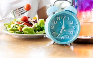 Jejum Intermitente: 5 Dicas Especiais Para Iniciantes - Uma pessoa comendo verduras e legumes com um relógio ao lado em cima da mesa.