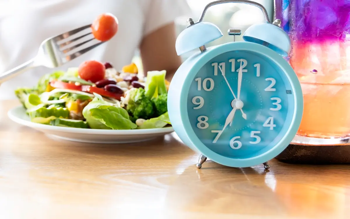 Jejum Intermitente: 5 Dicas Especiais Para Iniciantes - Uma pessoa comendo verduras e legumes com um relógio ao lado em cima da mesa.