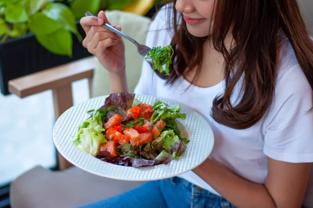 Mulher segurando um prato, levando salada com o garfo para a boca